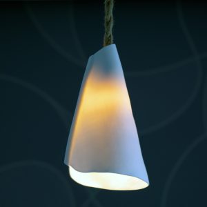 Suspension lumineuse en porcelaine blanche de forme organique, diffusant une lumière chaude, accrochée à un cordon en jute sur fond sombre.