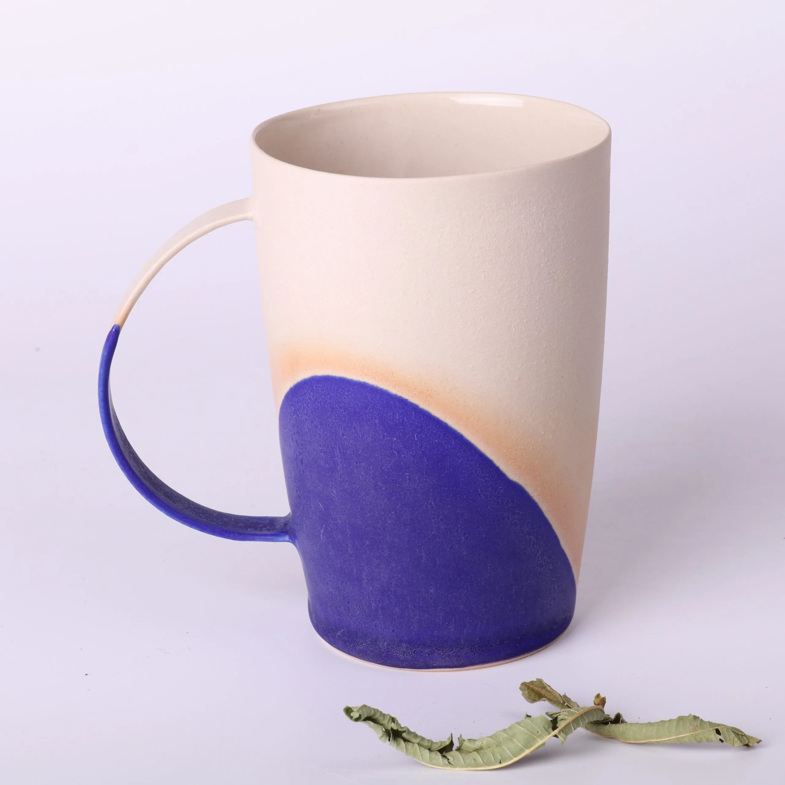 Mug poterie artisanale Bleu Klein — prêt pour le thé, 330 ml Mug poterie artisanale en grès avec émail Bleu Klein — 330 ml, anse fine ; feuilles de thé/verveine en avant-plan.