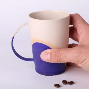 Mug poterie artisanale Bleu Klein, 330 ml — tenu en main pour l’échelle, base en grès naturel, grains de café au premier plan.