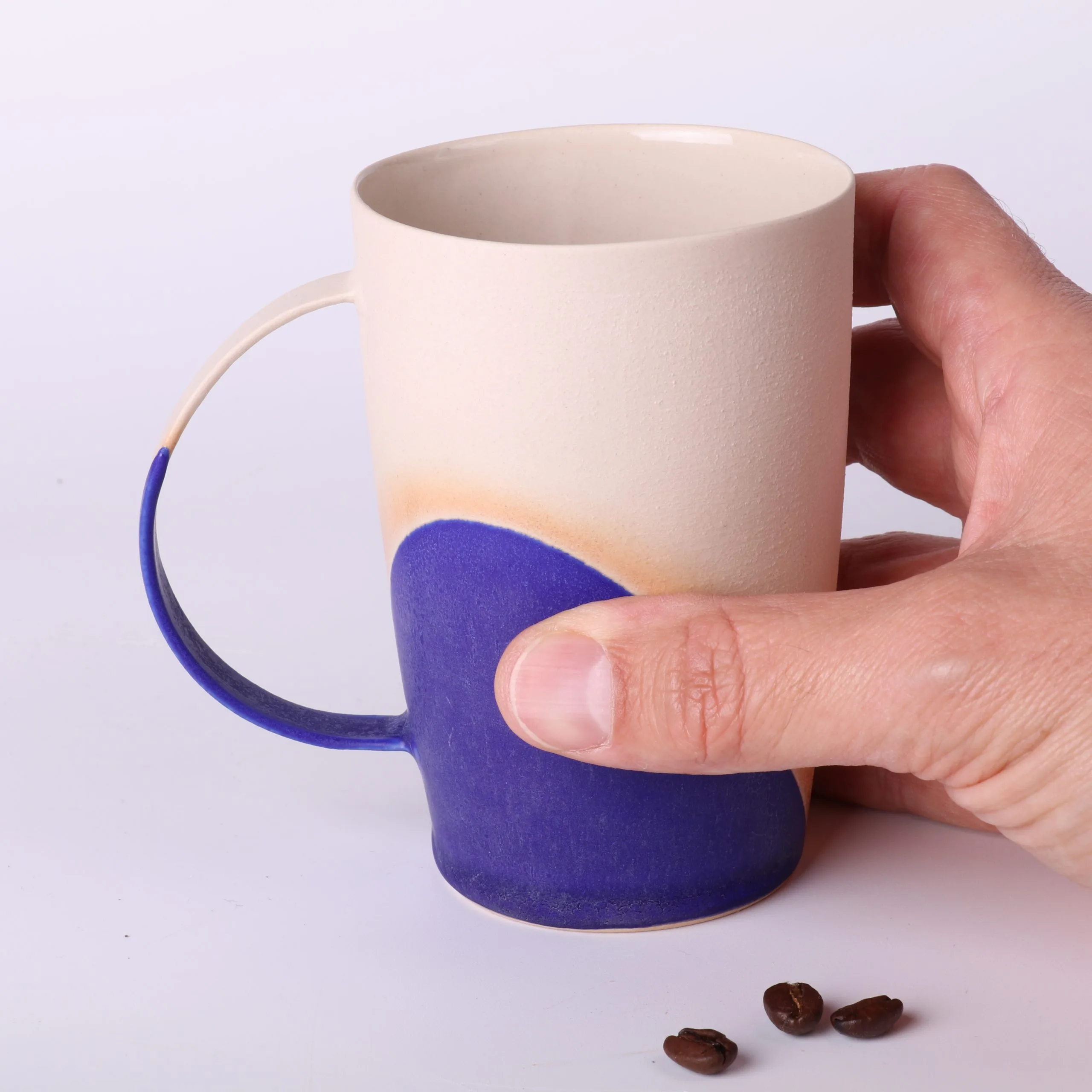 Mug poterie artisanale Bleu Klein — tenue en main, échelle réelle Mug poterie artisanale Bleu Klein, 330 ml — tenu en main pour l’échelle, base en grès naturel, grains de café au premier plan.