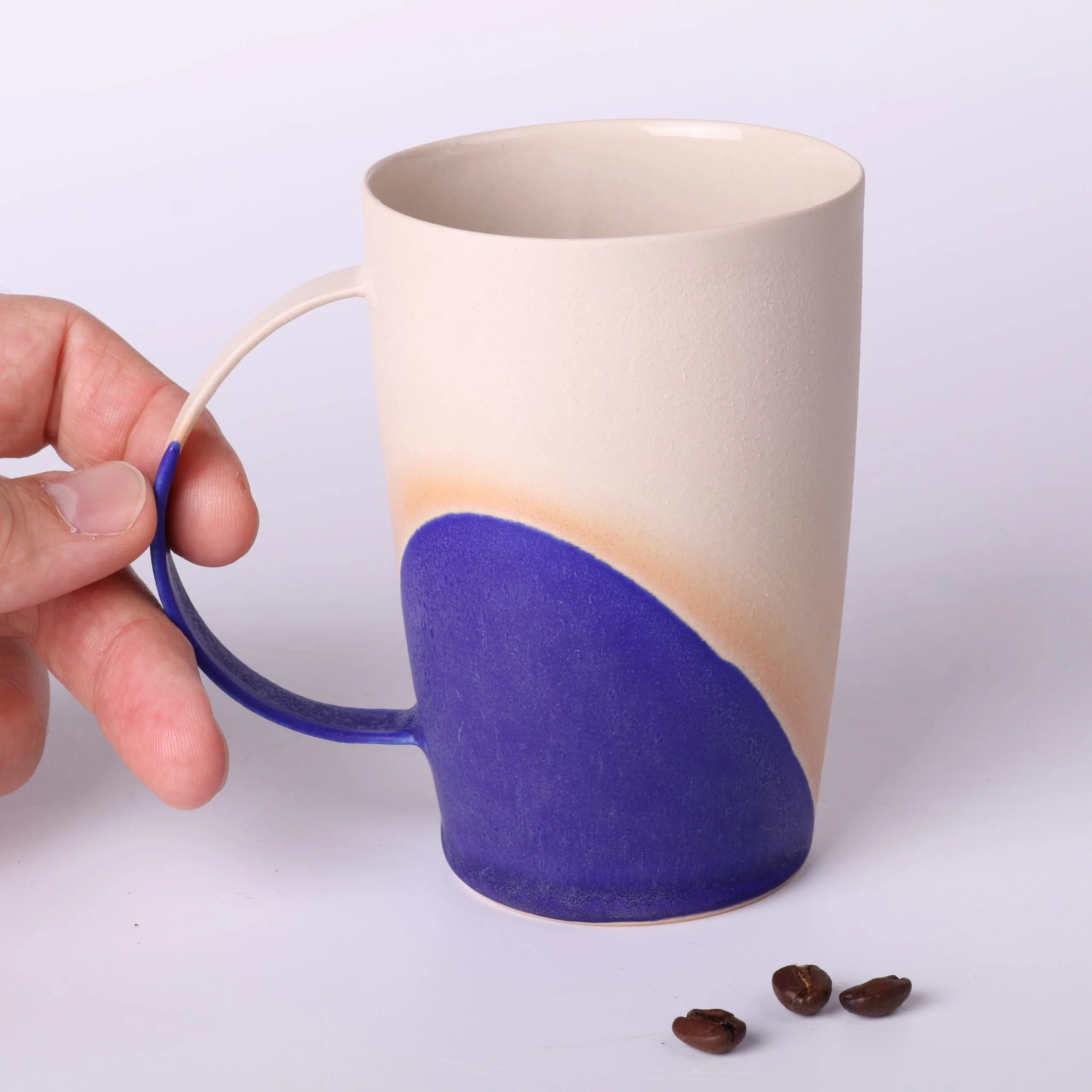 Mug poterie artisanale Bleu Klein — anse fine & grains de café Mug poterie artisanale Bleu Klein en grès, 330 ml — anse fine tenue du bout des doigts, trois grains de café au premier plan.