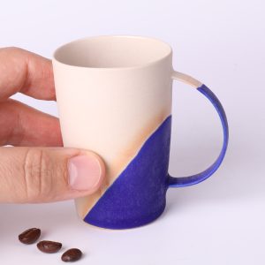 Tasse expresso design Bleu Klein tenue en main — échelle réelle, parois fines, grès crème et émail intense.
