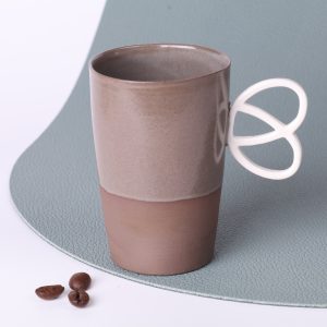 Tasse expresso en grès taupe 90 ml, faite main en Alsace, anse Bretzel ergonomique, vue trois-quarts sur fond vert sauge, grains de café au premier plan.