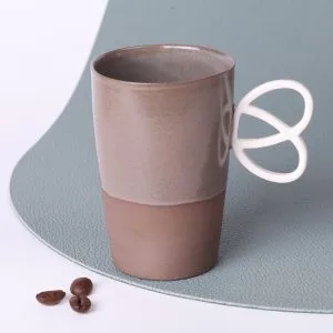 Tasse expresso en grès taupe 90 ml, faite main en Alsace, anse Bretzel ergonomique, vue trois-quarts sur fond vert sauge, grains de café au premier plan.