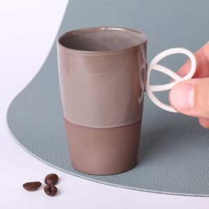 Tasse expresso anse bretzel Alsace 90 ml tenue entre les doigts, grès taupe vitrifié, vue rapprochée, grains de café visibles au premier plan.