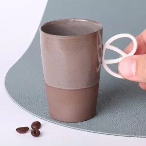 Tasse expresso anse bretzel Alsace 90 ml tenue entre les doigts, grès taupe vitrifié, vue rapprochée, grains de café visibles au premier plan.
