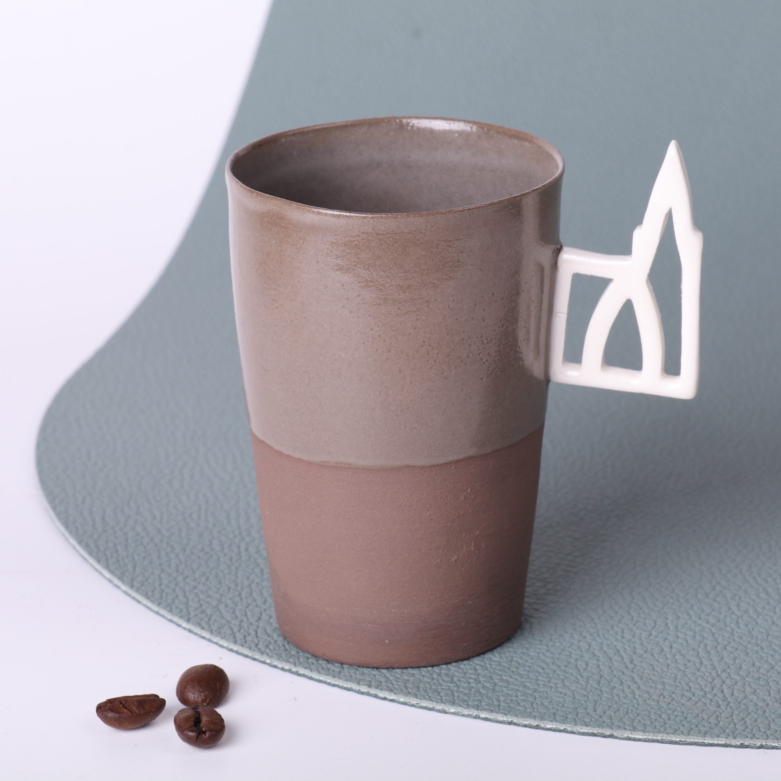 Tasse expresso design Alsace — grès taupe 90 ml, anse Cathédrale Tasse expresso en grès taupe 90 ml, faite main en Alsace, anse ajourée motif Cathédrale de Strasbourg en porcelaine, vue trois-quarts, grains de café au premier plan, fond vert sauge.