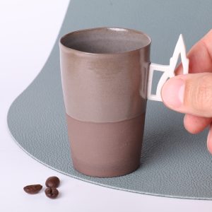 Tasse expresso design Alsace en grès taupe 90 ml tenue par l’anse ajourée motif Cathédrale de Strasbourg, vue rapprochée, grains de café au premier plan.