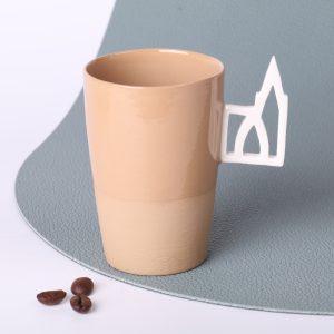 Tasse expresso en grès caramel 90 ml, faite main en Alsace, avec anse ajourée motif Cathédrale de Strasbourg en porcelaine, vue trois-quarts, grains de café au premier plan, fond vert sauge.