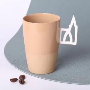 Tasse expresso en grès caramel 90 ml, faite main en Alsace, avec anse ajourée motif Cathédrale de Strasbourg en porcelaine, vue trois-quarts, grains de café au premier plan, fond vert sauge.