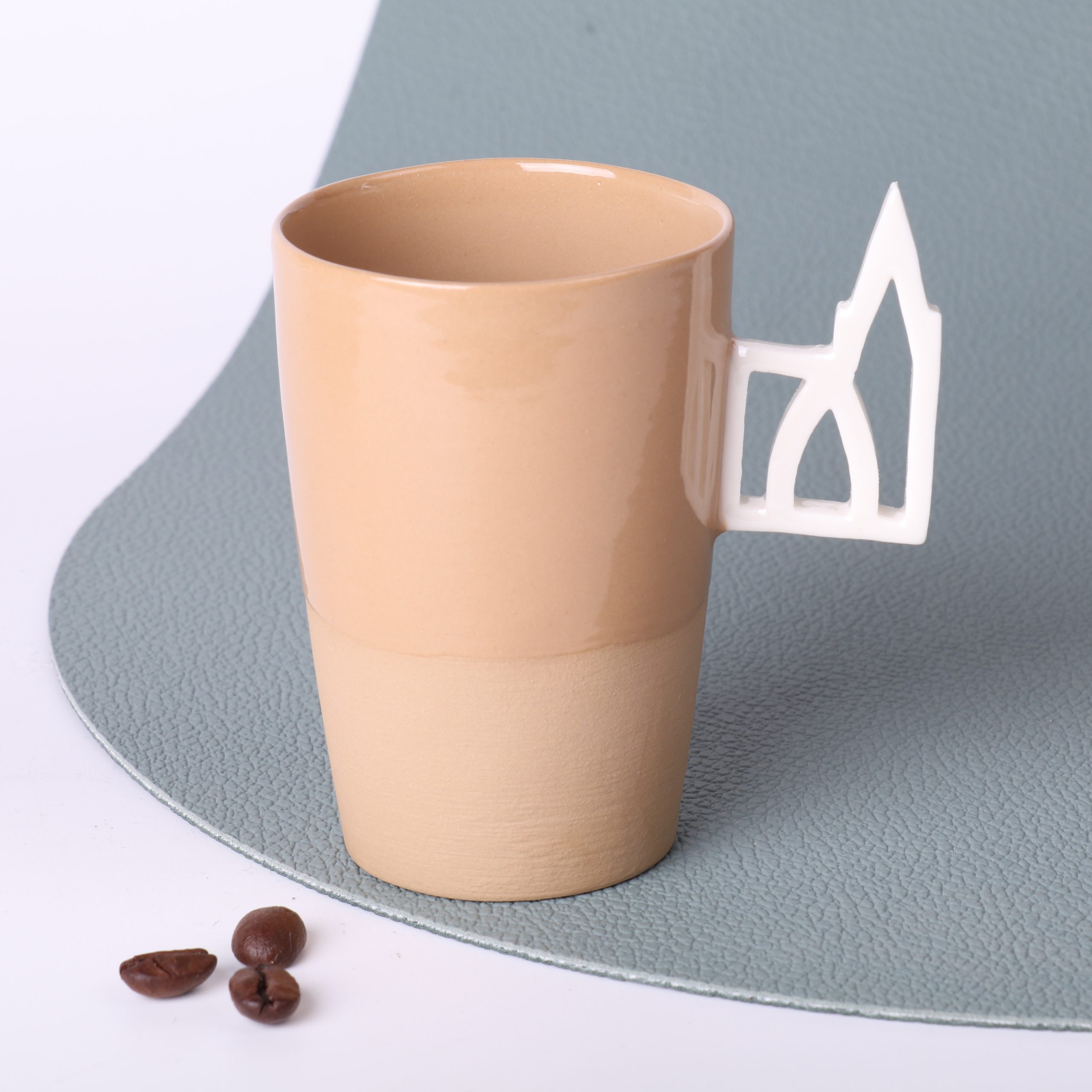 Tasse expresso caramel 90 ml – anse Cathédrale, grès fait main Alsace Tasse expresso en grès caramel 90 ml, faite main en Alsace, avec anse ajourée motif Cathédrale de Strasbourg en porcelaine, vue trois-quarts, grains de café au premier plan, fond vert sauge.