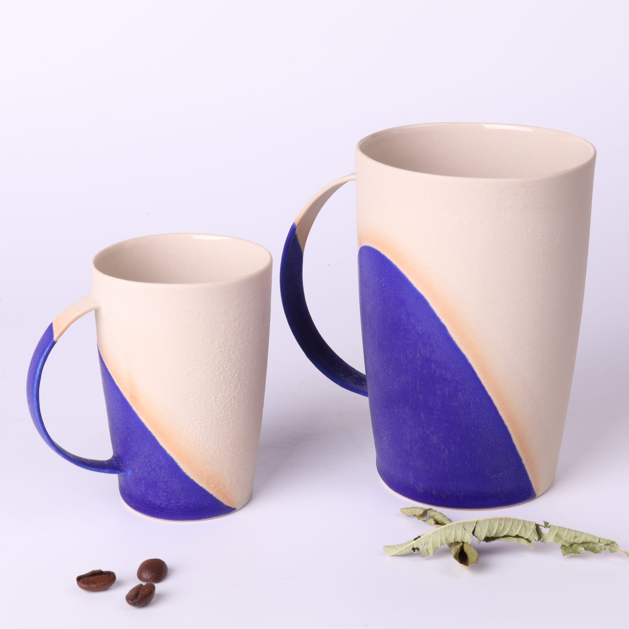mug-poterie-artisanale-bleu-klein-330ml-et-tasse-expresso-90ml Comparatif de taille : Mug poterie artisanale Bleu Klein 330 ml et tasse expresso design 90 ml en grès, côte à côte, avec grains de café et feuilles de verveine.