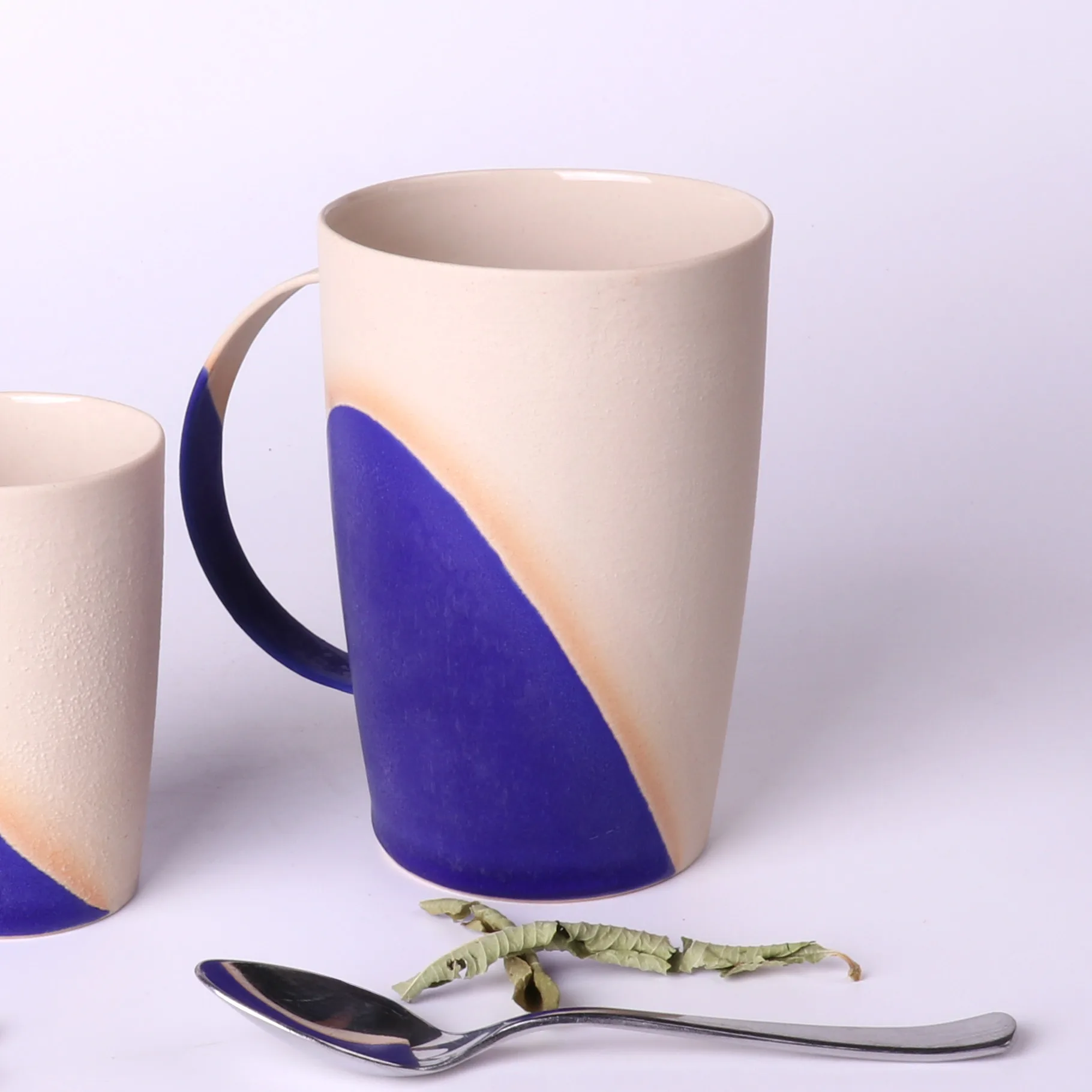 grand-mug-poterie-artisanale-bleu-klein-330ml-verveine-cuillere Grand mug poterie artisanale Bleu Klein 330 ml en grès, vue trois-quarts, avec cuillère inox et feuilles de verveine au premier plan; tasse expresso 90ml visible en bord gauche.