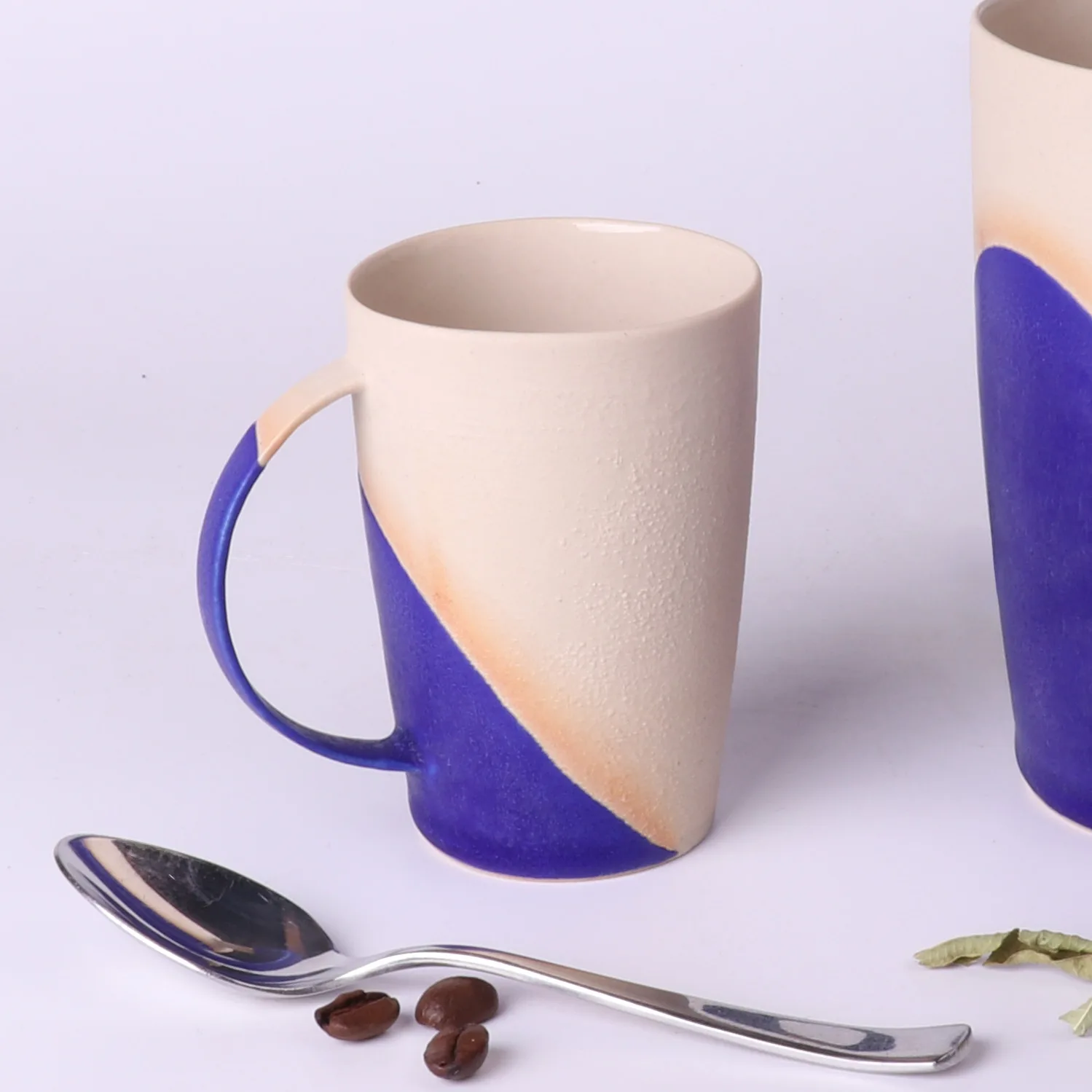 Tasse-expresso-poterie-artisanale-bleu-klein-90ml-cuillere-grains-cafe Tasse a expresso poterie artisanale Bleu Klein 90ml en grès, vue de trois-quarts avec cuillère à café inox et grains de café au premier plan; second mug 330ml en bord de cadre.