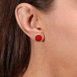 Boucle d’oreille puce ronde avec cabochon rouge brillant, portée à l’oreille d’une femme aux cheveux bruns.
